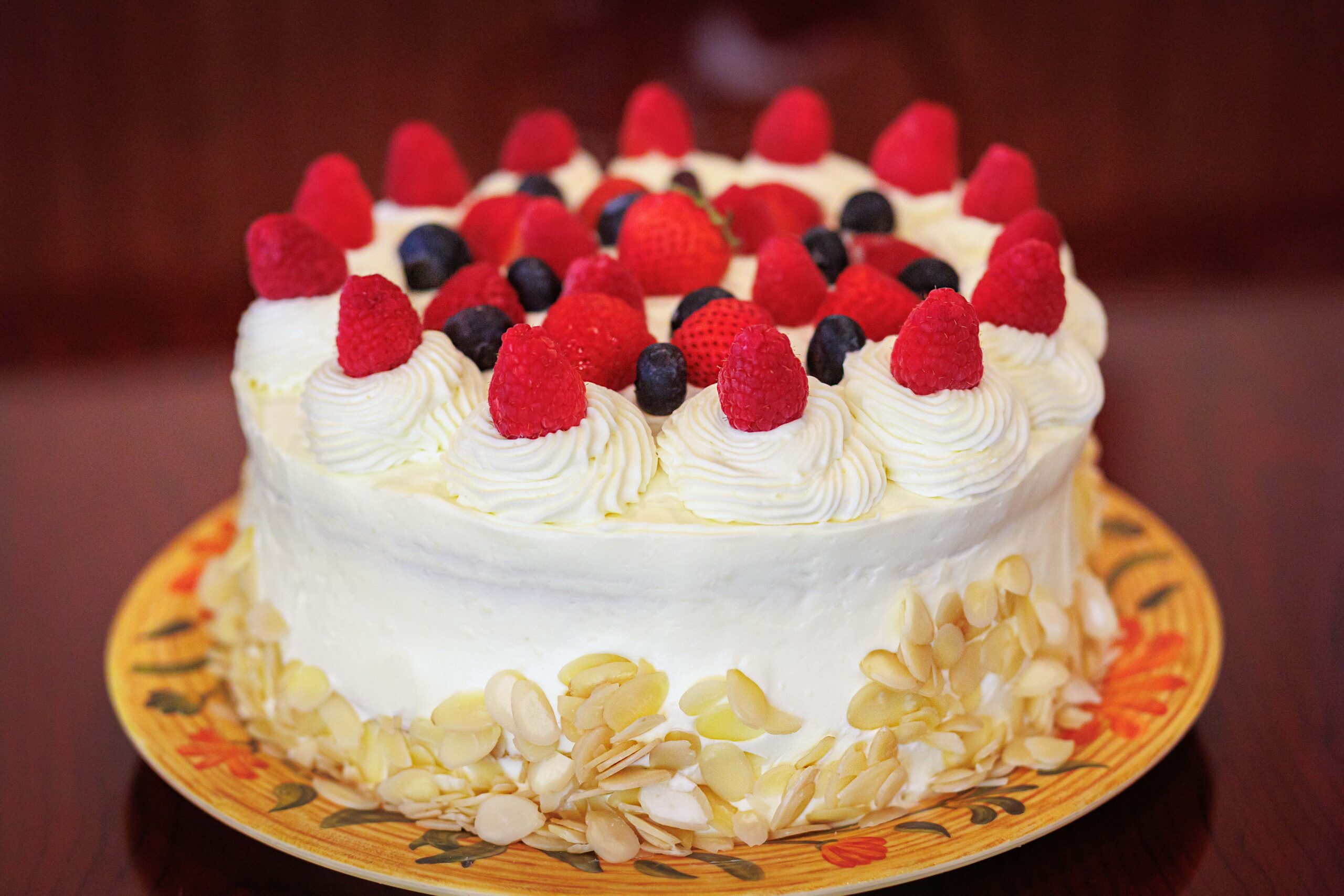 Round white-frosted cake topped with fresh raspberries and blueberries, with sliced almonds along the sides.