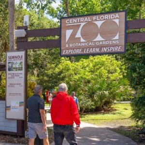 Entrance to Central Florida Zoo & Botanical Gardens with a large sign reading 'Explore. Learn. Inspire.' as people walk along a shaded path beneath trees.
