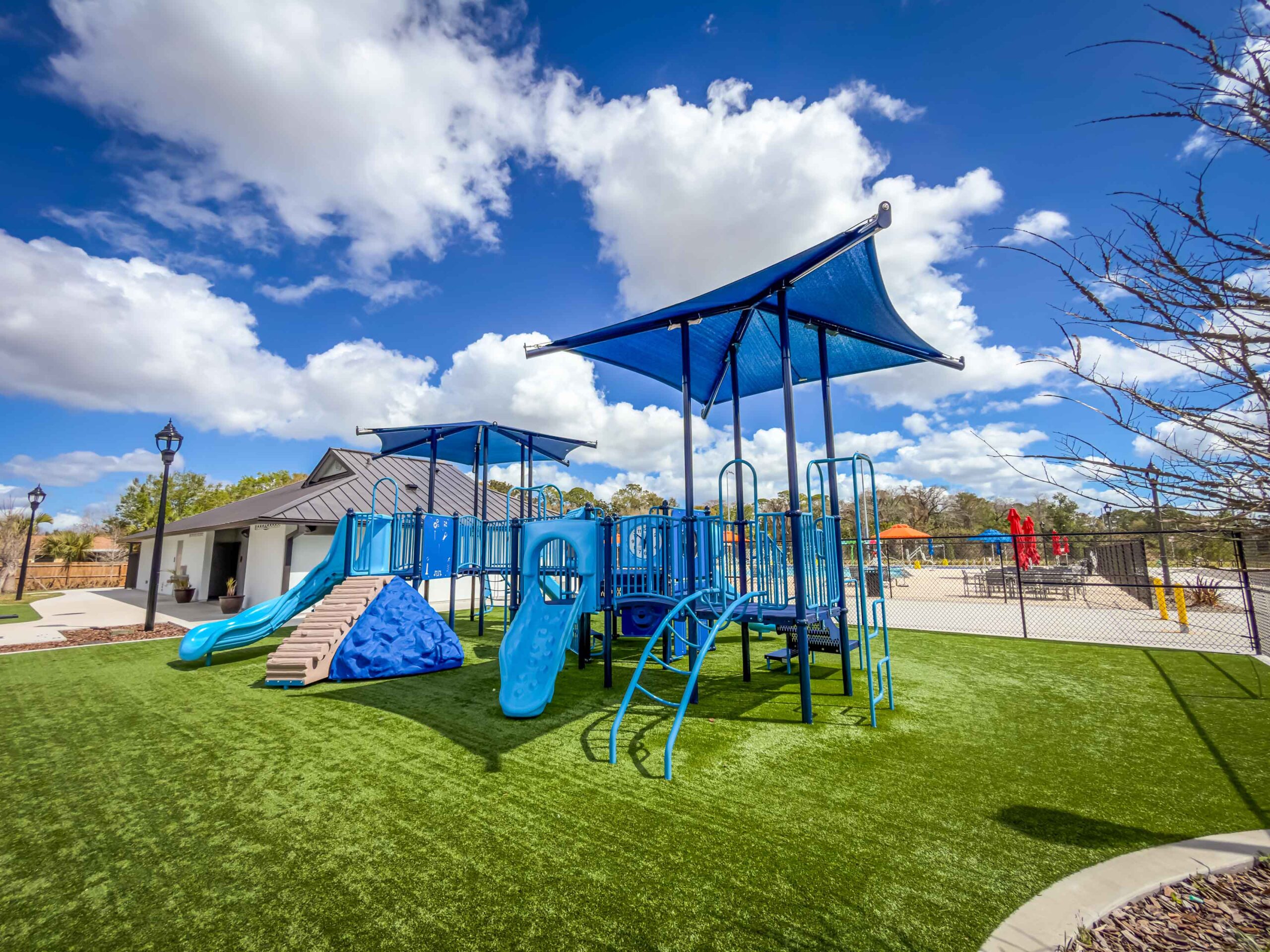Blue playground structures with slides and climbing elements on green artificial turf under a sunny blue sky.