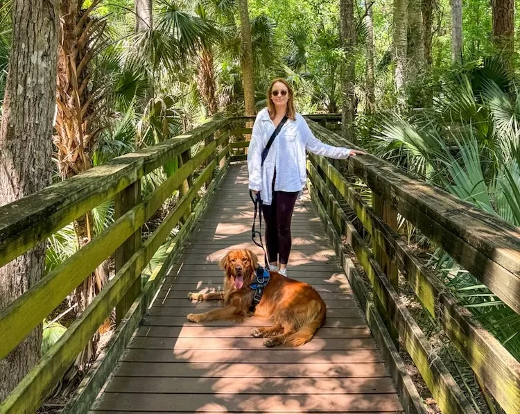 Woman walking a brown dog on a wooden boardwalk through a palm-filled jungle. She wears a white shirt and sunglasses, leash in hand.