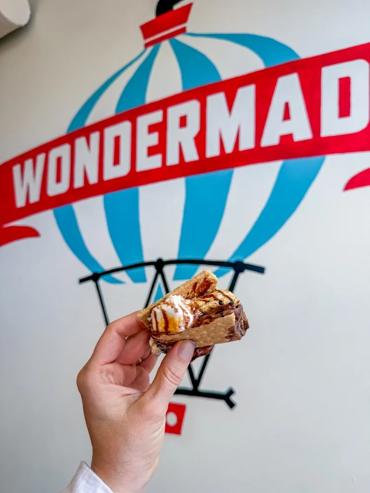 Hand holding a caramel-drizzled pastry in front of a red WONDERMAD mural with a blue balloon graphic.