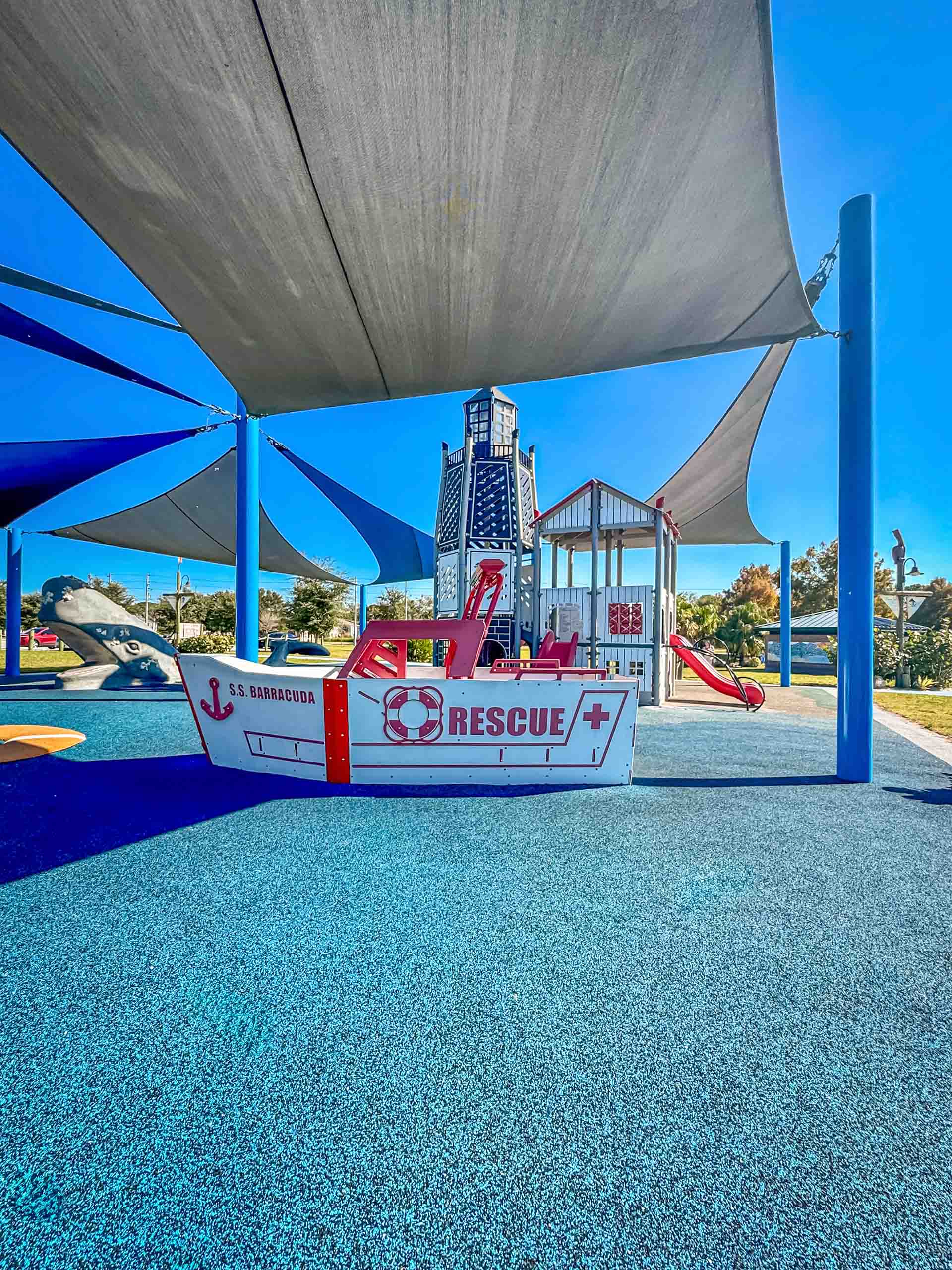 Playground under large blue shade sails with a white, boat-shaped play structure labeled RESCUE on a blue rubber surface.