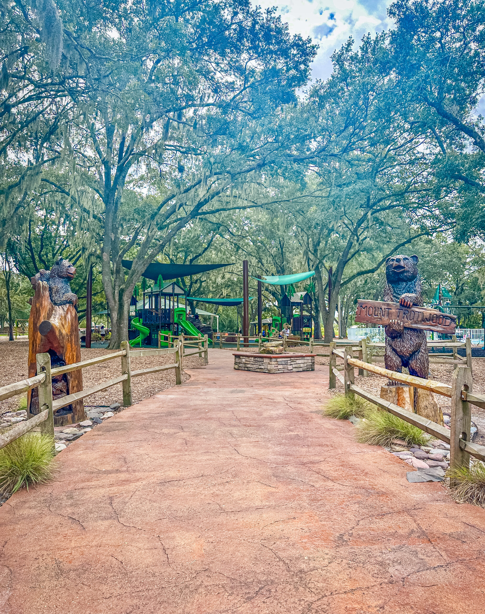 Park playground entrance with wooden bear sculptures on both sides of a cracked brick path under a canopy of trees, in a shaded outdoor area.